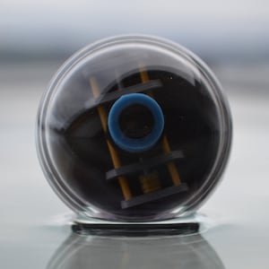 May include: A transparent, spherical object with a black interior, a blue ring, and yellow and black supports. The object is partially submerged in water, reflecting the sky above.