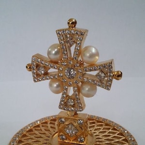 Może przedstawiać: Złoty krzyż z ozdobnym wzorem i białymi perłami, stojący na złotej podstawie z okrągłym wzorem.