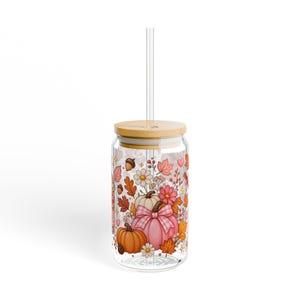 Puede incluir: Un vaso de vidrio transparente con tapa de bambú y pajita transparente, que presenta un estampado envolvente de calabazas naranjas y rosas, hojas de otoño, pequeñas flores blancas, bellotas y diminutos corazones rosas sobre fondo transparente.