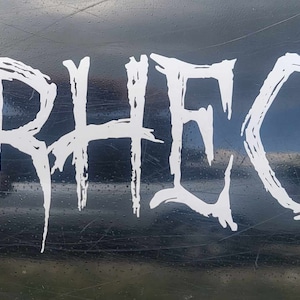 Puede incluir: Texto blanco estilizado que deletrea "RHEC" sobre un fondo negro. Las letras tienen un estilo de fuente gótica que gotea. El fondo es oscuro y reflectante, con arañazos visibles.