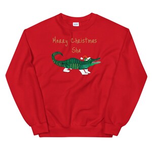 May include: Red Christmas sweatshirt with a green alligator wearing a Santa hat and the text "Merry Christmas Sha" in gold lettering.