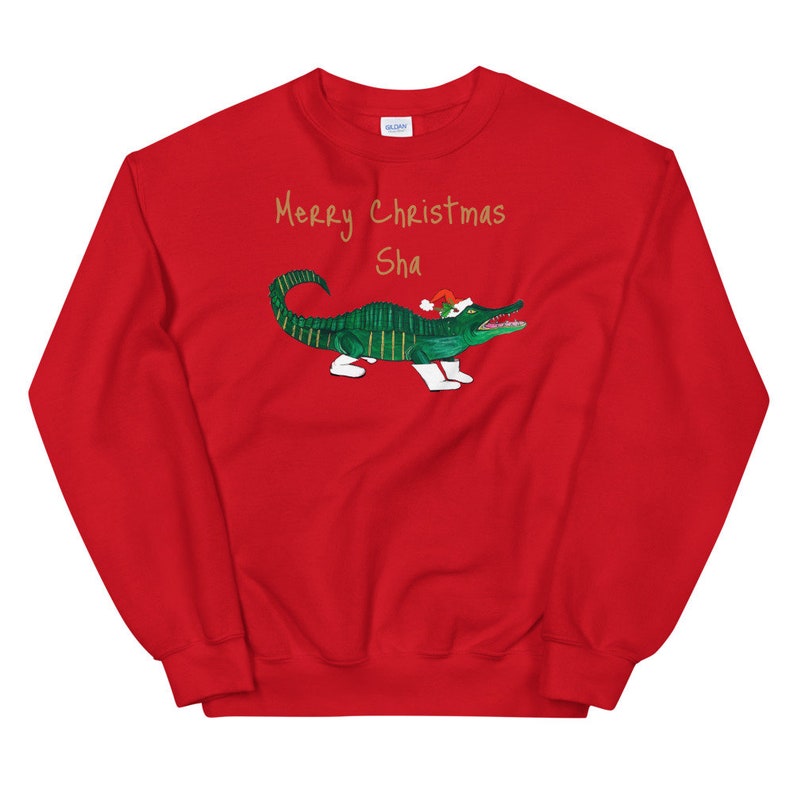 May include: Red Christmas sweatshirt with a green alligator wearing a Santa hat and the text "Merry Christmas Sha" in gold lettering.