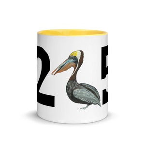 Puede incluir: Taza de cerámica blanca con borde amarillo. La taza presenta un número negro "2" en el lado izquierdo y un número negro "5" en el lado derecho. Un pelícano marrón y gris con un pico amarillo está centrado entre los números.