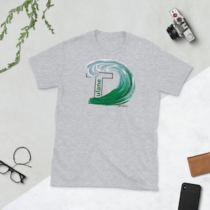 May include: A gray t-shirt with a green and blue wave design. The wave is stylized to look like the letter "T" and the word "Tulane" is written vertically inside the wave.