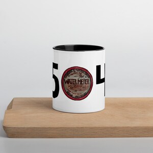 Könnte beinhalten: Weiße Keramiktasse mit schwarzem Rand. Die Tasse hat ein schwarzes "5" und "4" auf beiden Seiten eines roten und weißen runden Aufklebers mit der Aufschrift "Crescent City Water Meter Co."