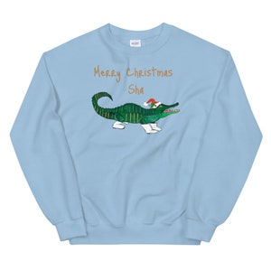 May include: Light blue sweatshirt with a green alligator wearing a Santa hat and white boots. The text "Merry Christmas Sha" is printed in orange on the sweatshirt.