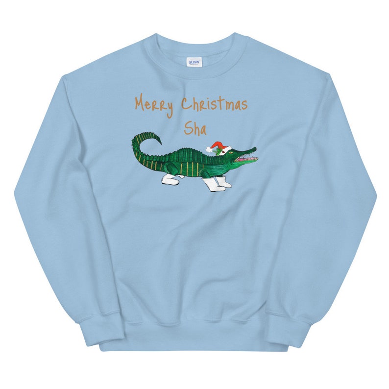 May include: Light blue sweatshirt with a green alligator wearing a Santa hat and white boots. The text "Merry Christmas Sha" is printed in orange on the sweatshirt.
