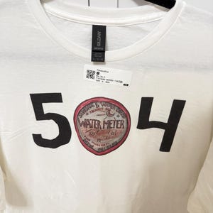 Op de afbeelding: Wit T-shirt met ronde hals met de zwarte cijfers "504" die een cirkelvormige afbeelding omlijsten. De afbeelding toont "Water Meter" met extra tekst en illustraties in rood en bruin.