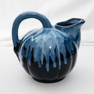 May include: A ceramic jug with a rounded body and a large handle. The jug is glazed in shades of blue and black, with a dripped effect. The spout is on the top, and the jug is sitting on a white surface.