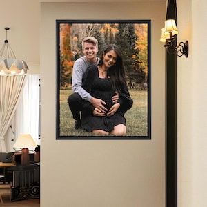 May include: A framed photograph featuring a couple posing outdoors. The woman, in a black dress, is cradling her pregnant belly. The man, in a gray long-sleeve shirt, has his arms around her. The background shows a forest with autumn colors.