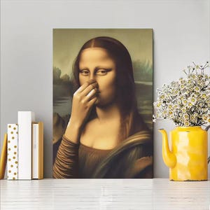 May include: A canvas print of the Mona Lisa, with the subject pinching her nose. The artwork is displayed on a white surface, next to a yellow teapot vase filled with white flowers. Books are on the left.