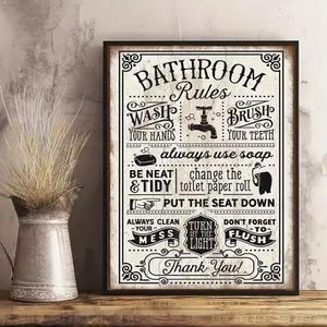 May include: A framed bathroom rules sign with black text on a white background. The sign includes instructions like "Wash Your Hands" and "Brush Your Teeth." A decorative metal pitcher with dried grass sits to the left.