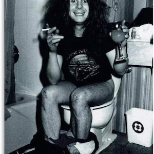 May include: Black and white photograph of a person sitting on a toilet, holding a lit cigarette and a drink. The person is wearing a black t-shirt with a band logo. The image has a vintage, candid feel.