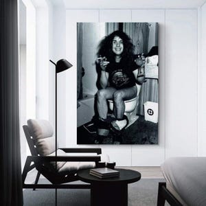 May include: Black and white framed print of a person sitting on a toilet, holding a cigarette and a drink. The person is wearing a t-shirt. A trash can is visible next to the toilet. The print is displayed in a modern room setting.