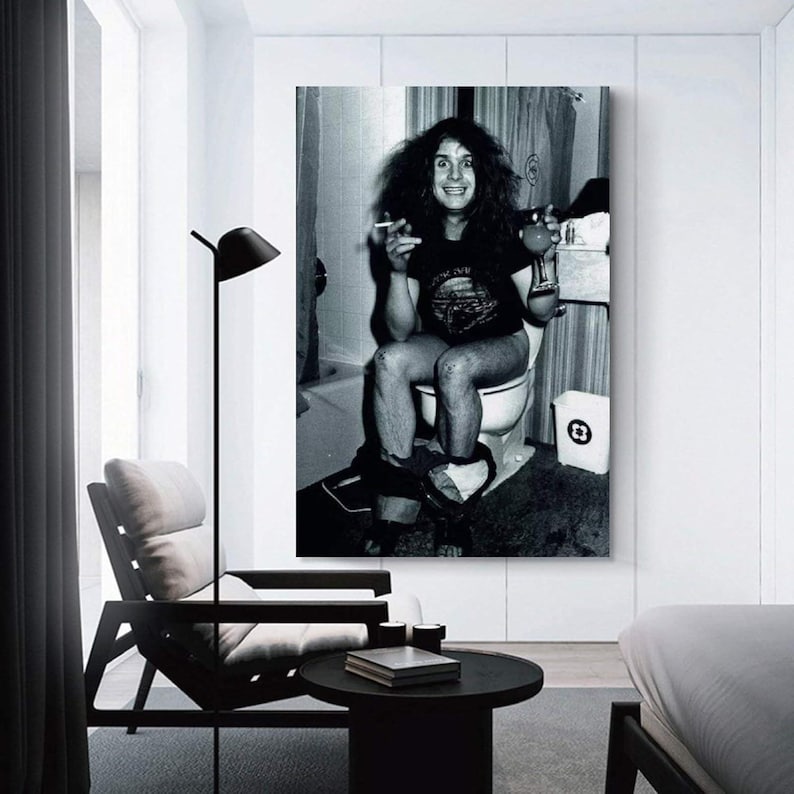 May include: Black and white framed print of a person sitting on a toilet, holding a cigarette and a drink. The person is wearing a t-shirt. A trash can is visible next to the toilet. The print is displayed in a modern room setting.