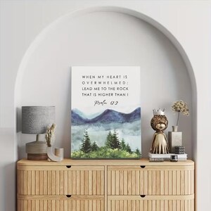 May include: A framed canvas print featuring the text "WHEN MY HEART IS OVERWHELMED; LEAD ME TO THE ROCK THAT IS HIGHER THAN I Psalm 61:2" above a watercolour landscape of mountains and trees. The print is displayed on a wooden cabinet.