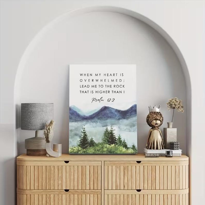May include: A framed canvas print featuring the text "WHEN MY HEART IS OVERWHELMED; LEAD ME TO THE ROCK THAT IS HIGHER THAN I Psalm 61:2" above a watercolour landscape of mountains and trees. The print is displayed on a wooden cabinet.