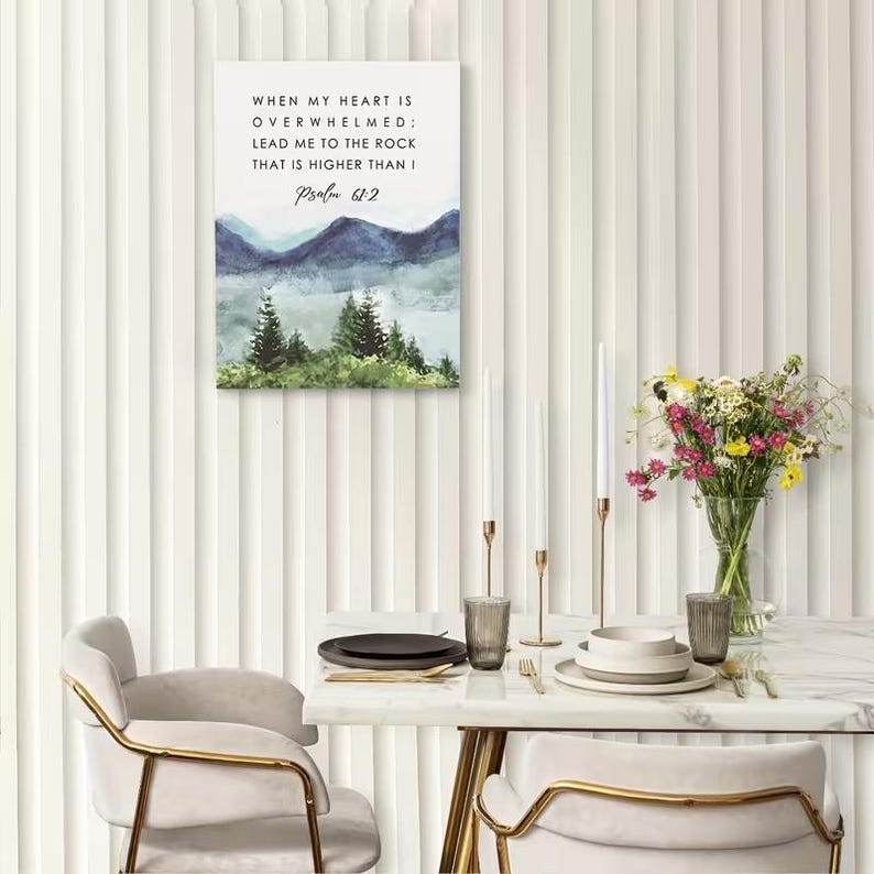 May include: A framed canvas print with the text "WHEN MY HEART IS OVERWHELMED; LEAD ME TO THE ROCK THAT IS HIGHER THAN I Psalm 61:2". The artwork features a watercolor landscape of mountains and trees. The scene is set on a table with a vase of flowers.