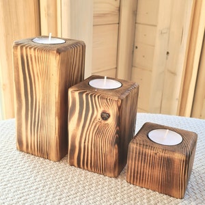 May include: Three wooden candle holders of varying sizes, each with a white tea light candle inside. The holders are made of rustic, dark brown wood with a natural wood grain pattern.