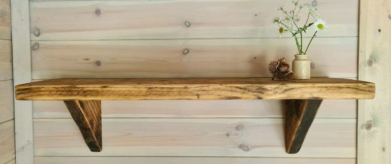 May include: A rustic wooden shelf with a natural wood finish and angled brackets. The shelf is mounted on a light wood wall and has a small vase with white flowers on top.