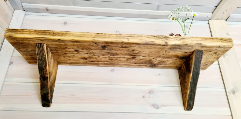 May include: A rustic wooden shelf with a natural wood finish and angled brackets. The shelf is mounted on a white wall.