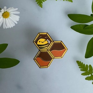 May include: A gold-colored enamel pin featuring a cartoon bee inside a honeycomb. The bee is yellow with black stripes and has a smiling face. The honeycomb is brown and orange.