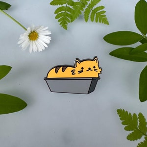 Puede incluir: Un gato amarillo y marrón está durmiendo en un molde de pan gris. El gato es un pin de esmalte de estilo de dibujos animados.