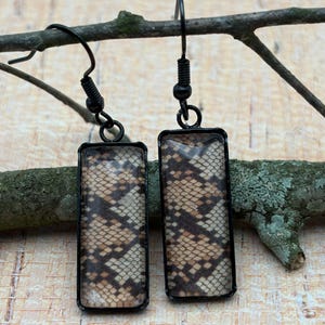 May include: A pair of rectangular drop earrings featuring a black metal frame and a brown and beige snakeskin pattern inlay. The earrings are attached to black metal fishhook wires, providing a simple and clean design for these dangle earrings.