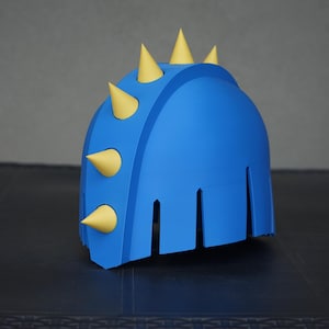 Puede incluir: Un casco azul brillante con una cresta central adornada con cuatro púas amarillas puntiagudas. El casco tiene un acabado mate suave y una serie de recortes verticales a lo largo del borde inferior, creando una apariencia segmentada.