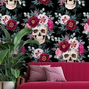 May include: A black background with a repeating pattern of white skulls wearing floral crowns of pink and burgundy flowers. The skulls are surrounded by green leaves. A red sofa sits in front of the wallpaper.