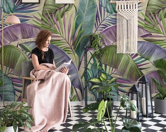 Op de afbeelding: Een vrouw zit op een stoel en leest een boek in een kamer met een tropische bladbehang, een roze bank en verschillende planten in potten.