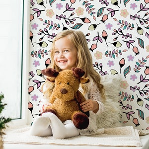 Puede incluir: Una niña pequeña se sienta en un asiento de ventana con un juguete de peluche de reno marrón. La pared detrás de ella está cubierta con un papel tapiz blanco con un patrón floral rosa, verde y azul.