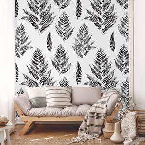 K&ouml;nnte beinhalten: Ein Wohnzimmer-Interieur mit einer wei&szlig;en Wand, die mit einer schwarz-wei&szlig;en Farn-Mustertapete versehen ist. Vor der Wand steht ein hellgraues Sofa mit Kissen und einer Decke. Im Vordergrund steht ein kleiner Holztisch mit einer Schale voller Dekoartikel.