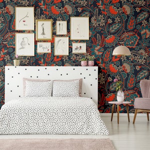 May include: A bedroom with a white bed with black polka dots bedding and a pink armchair. The wall is covered in a dark blue wallpaper with a floral pattern in orange, teal, and brown. There are framed pictures on the wall.