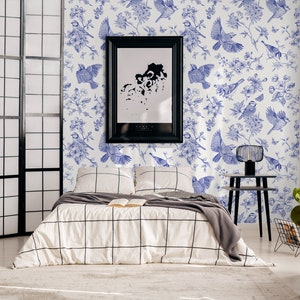 May include: A white bedroom with a black and white patterned bedspread. The wall is covered in a blue and white wallpaper with a floral and bird design. A black picture frame hangs on the wall above the bed. A black side table with a lamp sits next to the bed.