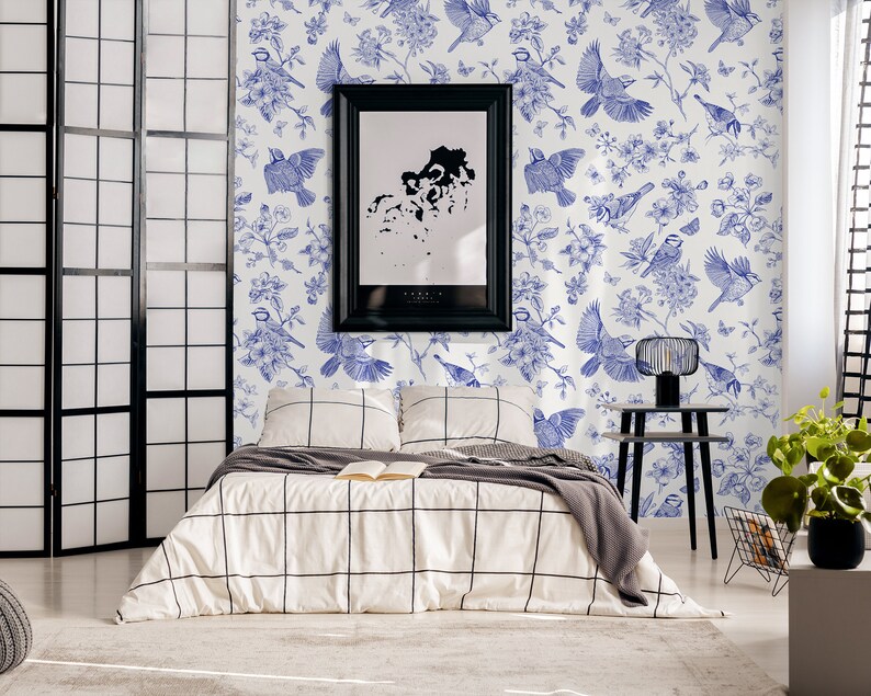 May include: A white bedroom with a black and white patterned bedspread. The wall is covered in a blue and white wallpaper with a floral and bird design. A black picture frame hangs on the wall above the bed. A black side table with a lamp sits next to the bed.
