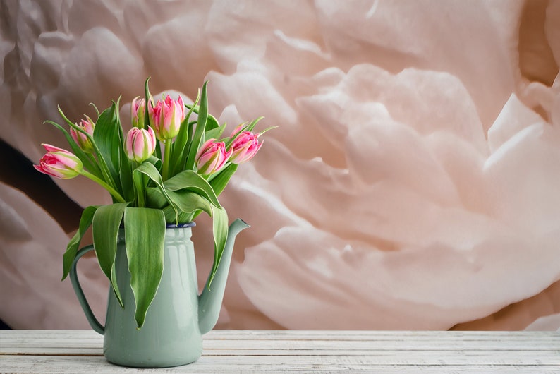 K&ouml;nnte beinhalten: Ein Strau&szlig; rosa und wei&szlig;er Tulpen in einem hellblauen Emaille-Krug auf einem wei&szlig;en Holztisch. Der Hintergrund ist ein weiches, pfirsichfarbenes abstraktes Design.