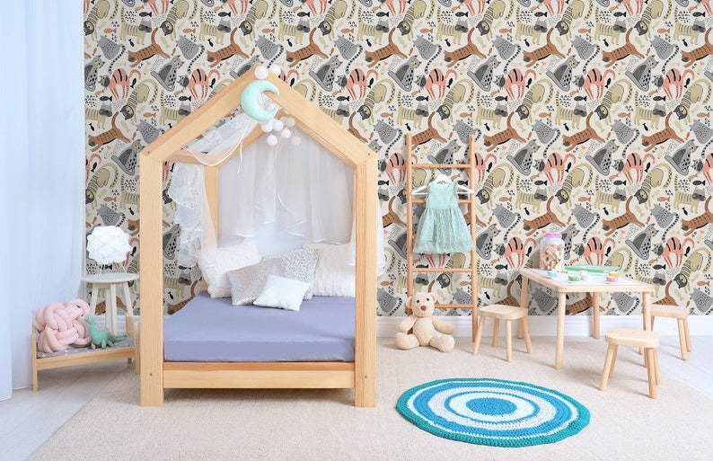 May include: A light blue canopy bed with a white canopy and a light blue rug. The wall behind the bed is covered in a wallpaper with a pattern of colorful cats and fish. There is a small wooden table with a white lamp on it, a wooden stool with a pink knitted toy on it, and a wooden ladder with a light blue dress hanging on it.