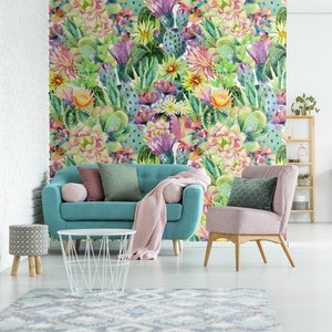 May include: A teal blue sofa with pink and green throw pillows sits in front of a wall covered in a watercolor cactus print wallpaper. A pink armchair with a green pillow sits to the right of the sofa. A white wire coffee table sits in front of the sofa. A small patterned stool sits to the left of the sofa.