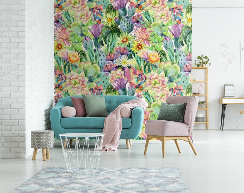 May include: A teal blue sofa with pink and green throw pillows sits in front of a wall covered in a watercolor cactus print wallpaper. A pink armchair with a green pillow sits to the right of the sofa. A white wire coffee table sits in front of the sofa. A small patterned stool sits to the left of the sofa.