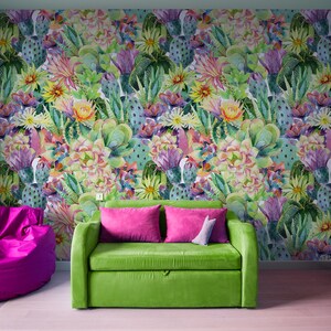 May include: A green sofa with pink pillows sits in front of a wall with a colorful cactus print wallpaper. A pink beanbag chair is on the left and a pink bookshelf is on the right.