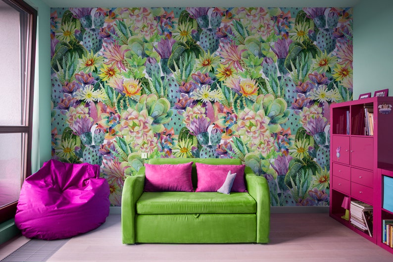 May include: A green sofa with pink pillows sits in front of a wall with a colorful cactus print wallpaper. A pink beanbag chair is on the left and a pink bookshelf is on the right.