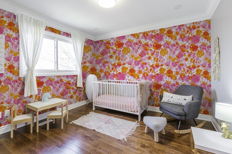 May include: A nursery room with a white crib, a rocking chair, a small table and chairs, and a rug. The walls are covered in a pink and orange splatter paint wallpaper.