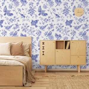 May include: A bedroom interior with a white wall featuring a blue and white wallpaper pattern of birds and flowers. A wooden bed with a white duvet and brown pillows is in the foreground. A wooden cabinet with shelves is to the right of the bed. A woven rug is on the floor.