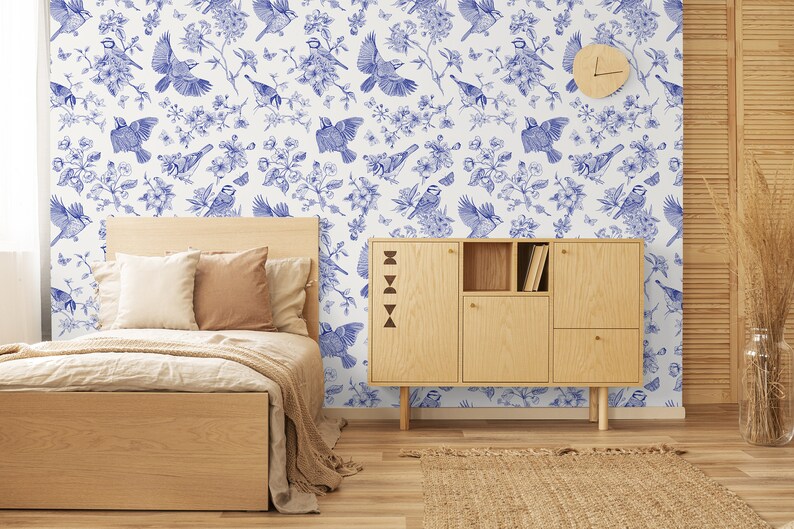 May include: A bedroom interior with a white wall featuring a blue and white wallpaper pattern of birds and flowers. A wooden bed with a white duvet and brown pillows is in the foreground. A wooden cabinet with shelves is to the right of the bed. A woven rug is on the floor.