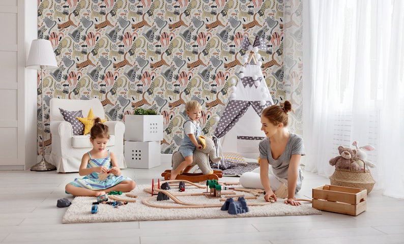 May include: A playroom with a white armchair, a teepee tent, and a wooden toy train set. Two children are playing with the train set, while a woman sits on the floor and watches them. The walls are covered in a colorful wallpaper with a pattern of animals.