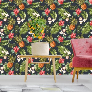May include: A dark blue wallpaper with a tropical print featuring pineapples, red flowers, and green leaves. A small potted citrus tree sits on a wooden table in front of the wallpaper, and a pink armchair is to the right of the table.