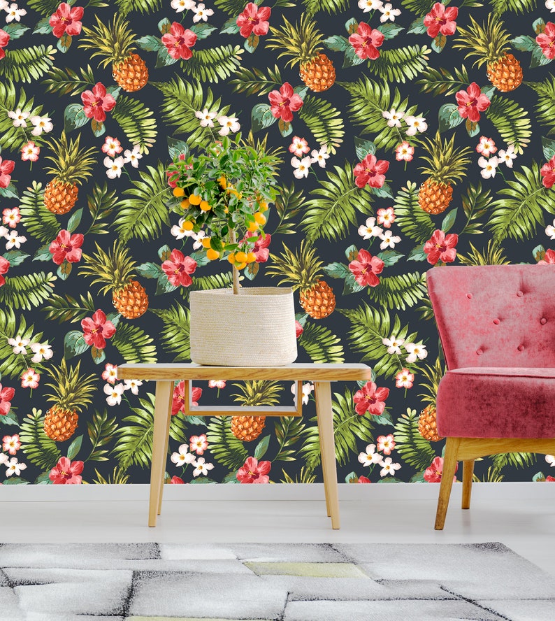 May include: A dark blue wallpaper with a tropical print featuring pineapples, red flowers, and green leaves. A small potted citrus tree sits on a wooden table in front of the wallpaper, and a pink armchair is to the right of the table.