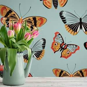 May include: A blue ceramic pitcher with a white rim, filled with pink tulips, sits on a white wooden surface in front of a blue wall with a repeating pattern of colorful butterflies.