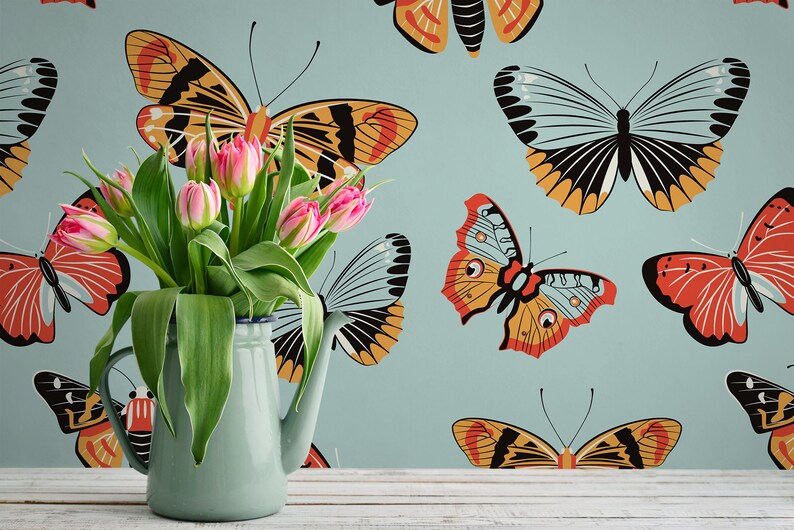 May include: A blue ceramic pitcher with a white rim, filled with pink tulips, sits on a white wooden surface in front of a blue wall with a repeating pattern of colorful butterflies.